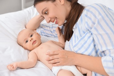 Happy young woman applying body cream onto baby`s skin on bed