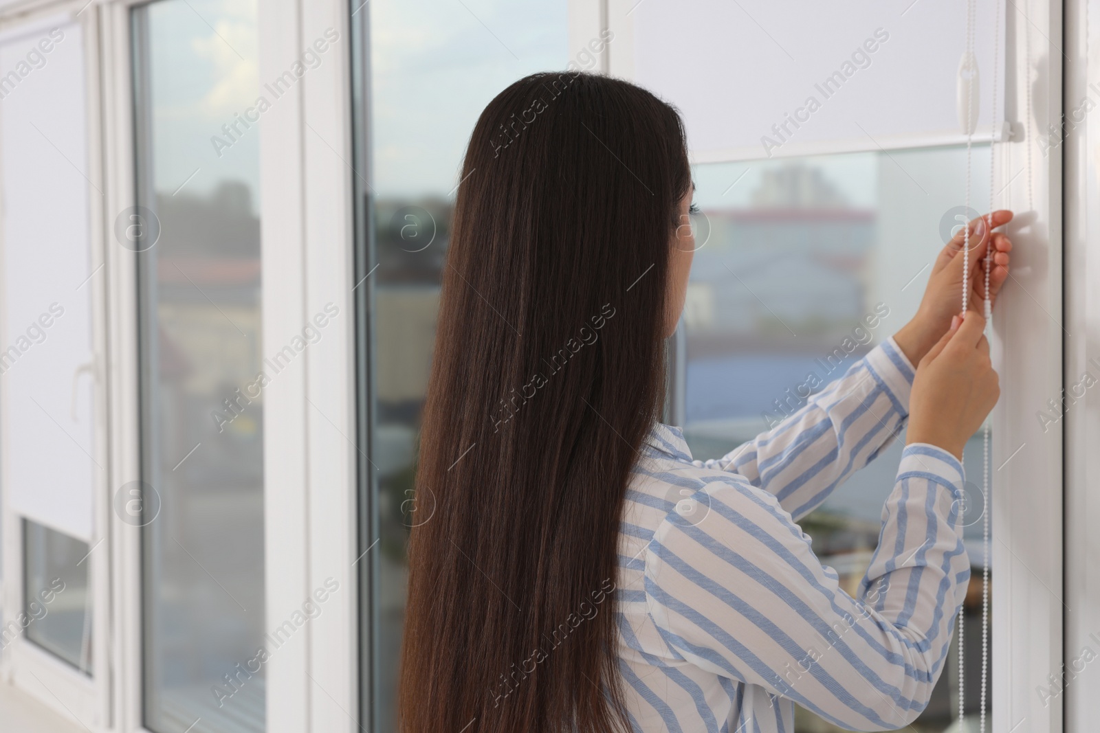 Photo of Woman opening white roller blinds on window indoors