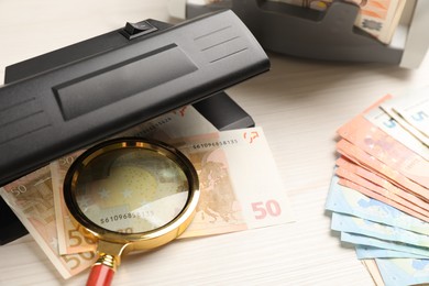 Photo of Modern currency detector with Euro banknotes and magnifying glass on white wooden table. Money examination device