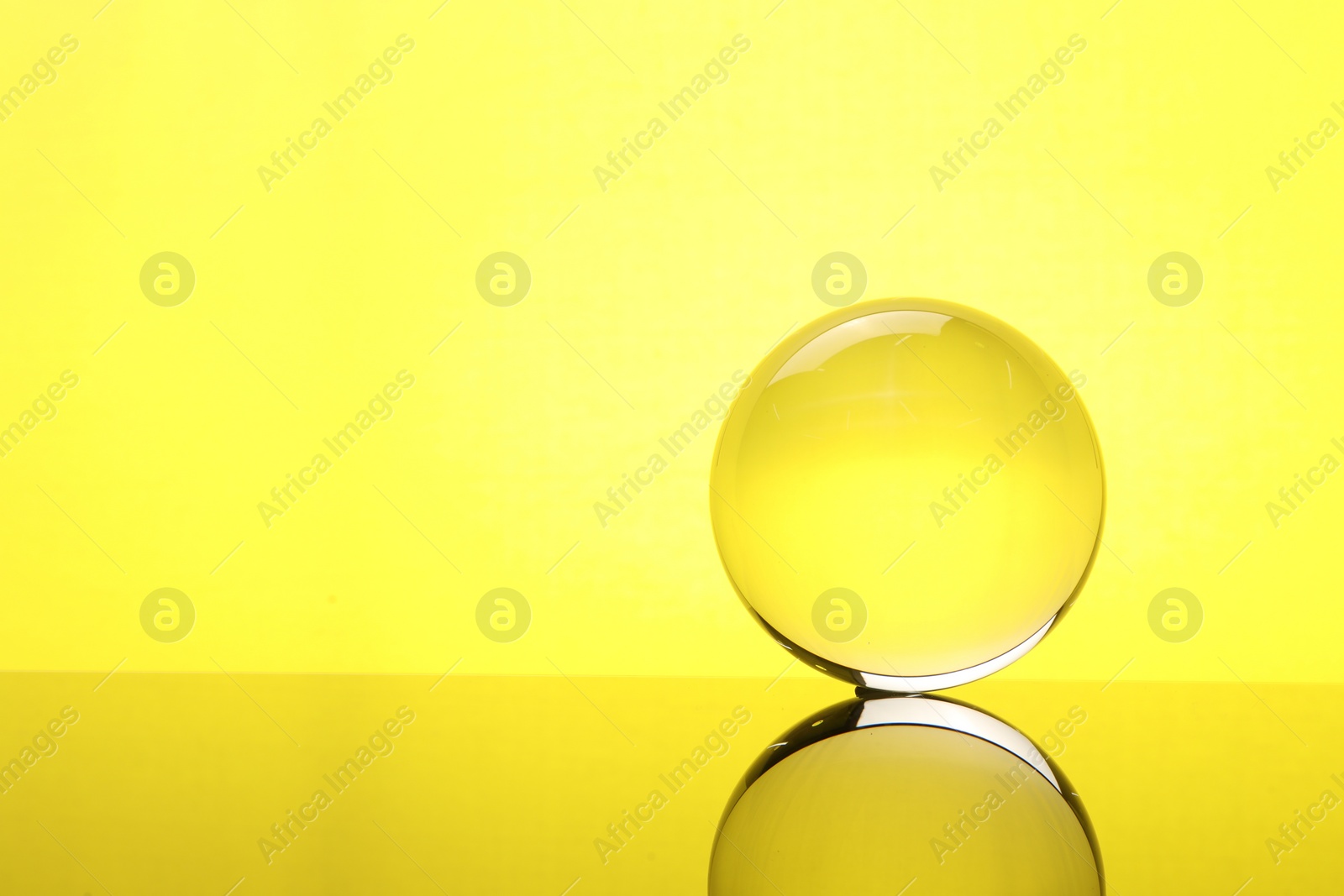 Photo of Transparent glass ball on mirror surface against yellow background. Space for text