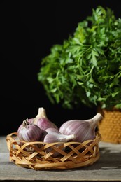 Photo of Fresh raw garlic in wicker basket and parsley on wooden table against black background