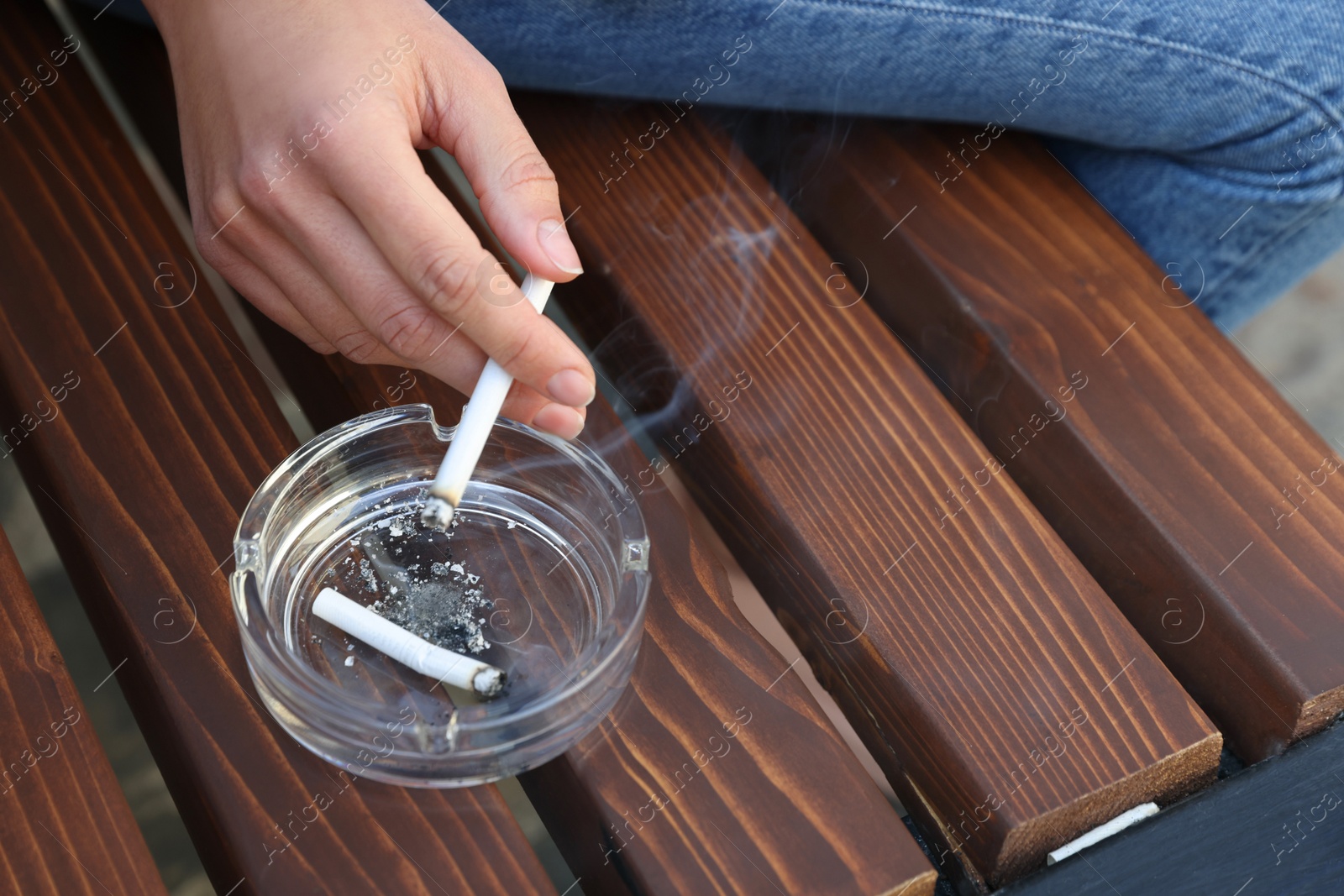 Photo of Woman holding cigarette over glass ashtray on bench outdoors, closeup