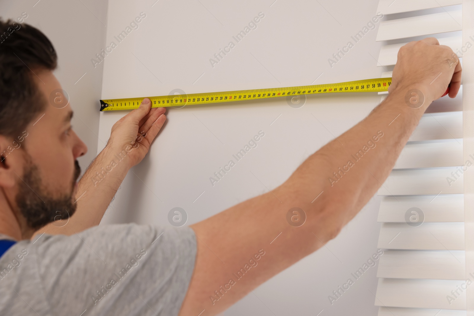 Photo of Worker in uniform using measuring tape while installing horizontal window blinds indoors