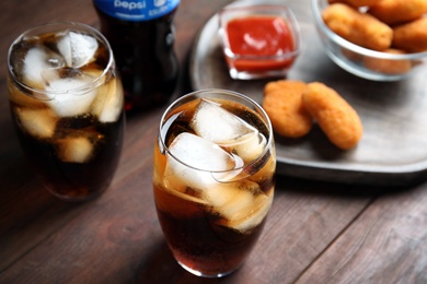 Photo of MYKOLAIV, UKRAINE - FEBRUARY 15, 2021: Glasses of Pepsi and snack on wooden table, closeup