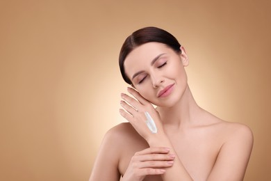 Beautiful woman with smear of body cream on her hand against light brown background, space for text