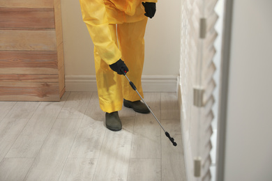 Photo of Pest control worker in protective suit spraying insecticide on floor indoors, closeup