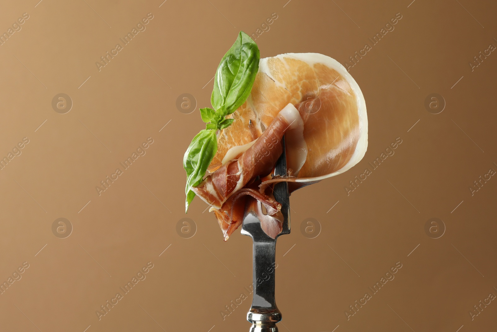 Photo of Fork with slice of tasty jamon and basil leaves on brown background, closeup