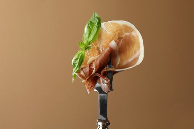 Fork with slice of tasty jamon and basil leaves on brown background, closeup