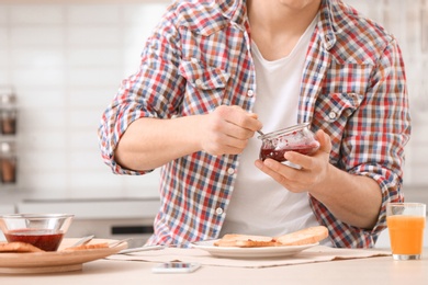 Photo of Young man with jar of jam and tasty toasted bread at table in kitchen