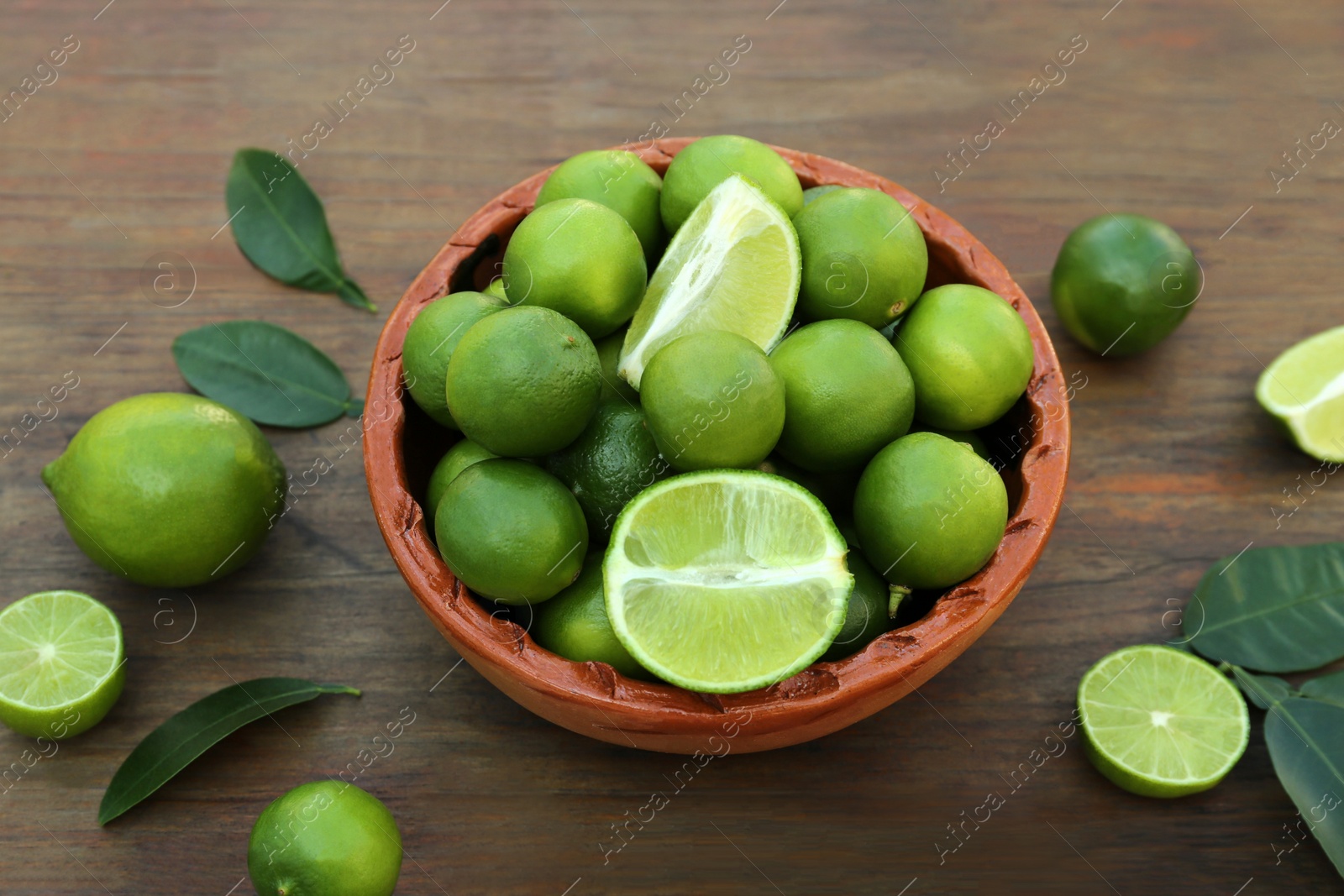 Photo of Whole and cut fresh ripe limes with green leaves in bowl on wooden table