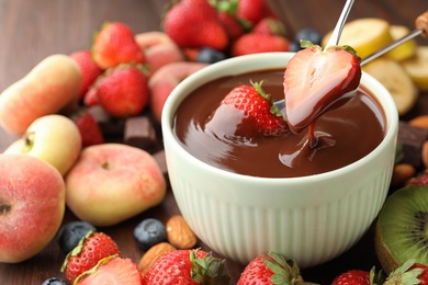 Photo of Fondue forks with strawberries in bowl of melted chocolate surrounded by other fruits on table