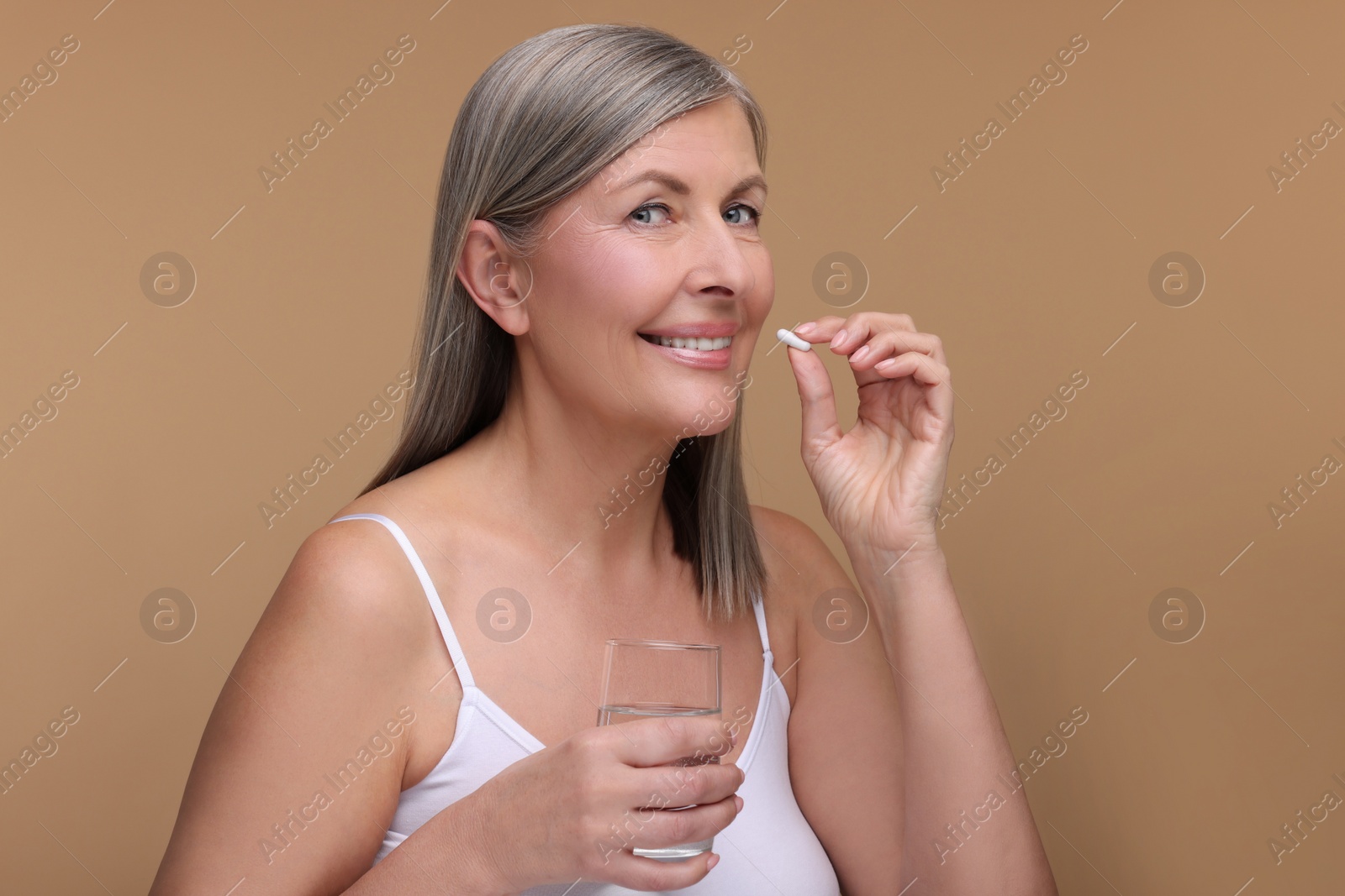 Photo of Beautiful woman with glass of water taking vitamin capsule on beige background