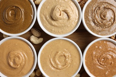 Photo of Many tasty nut butters in bowls and nuts on wooden table, flat lay