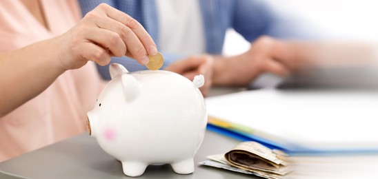 Image of Mature woman putting coin in piggy bank at table, closeup. Banner design