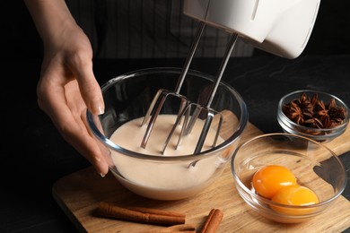Woman whipping ingredients with mixer at black table, closeup. Cooking delicious eggnog
