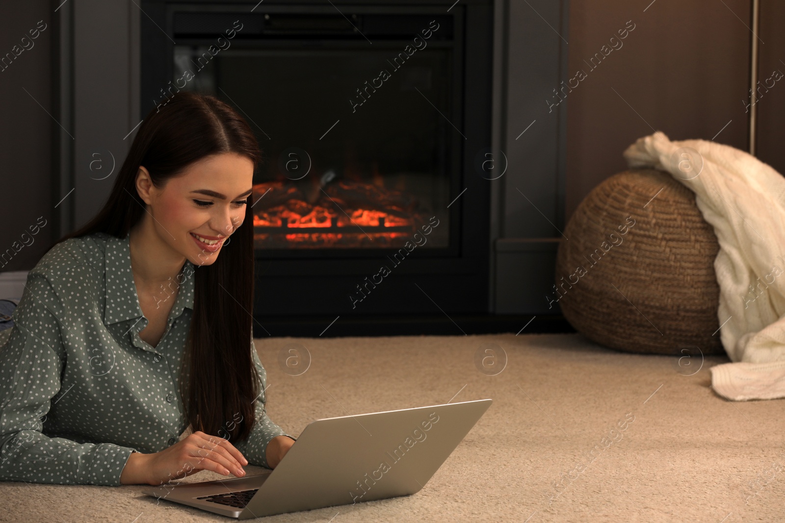 Photo of Beautiful young woman with laptop lying on floor near fireplace at home. Space for text