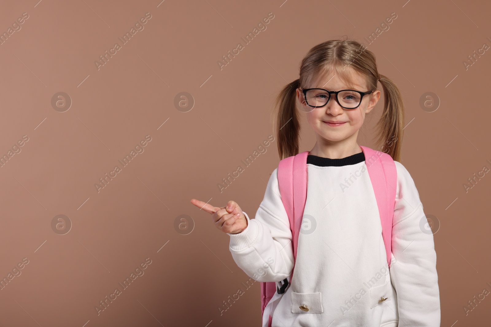 Photo of Happy schoolgirl in glasses with backpack pointing at something on brown background, space for text