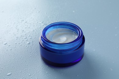 Moisturizing cream in open jar on light blue background with water drops, closeup