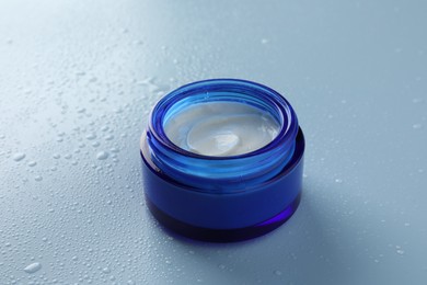 Photo of Moisturizing cream in open jar on light blue background with water drops, closeup