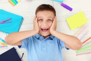 Photo of Emotional little boy with school stationery on wooden background, top view