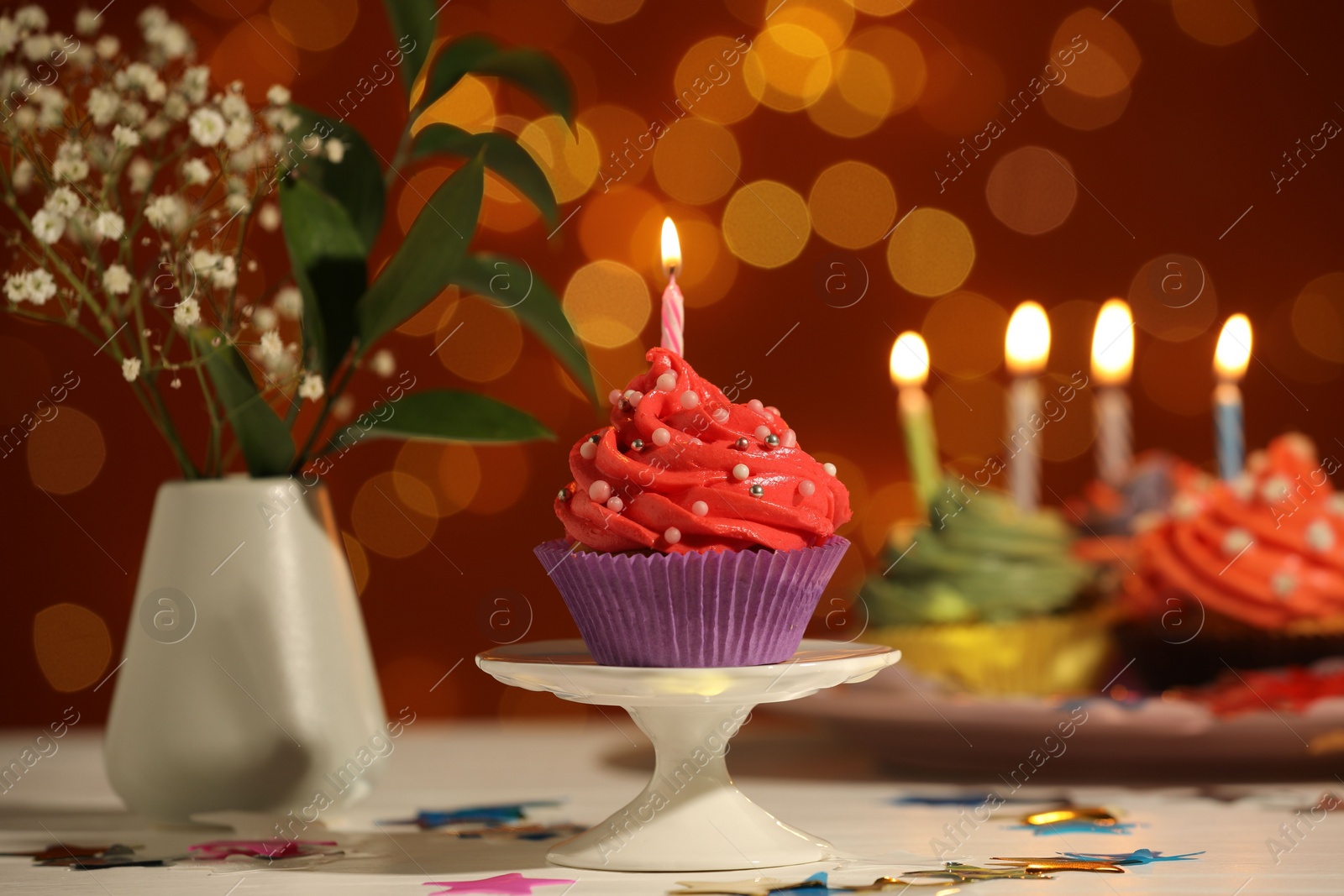 Photo of Stand with birthday cupcake on white table against blurred lights