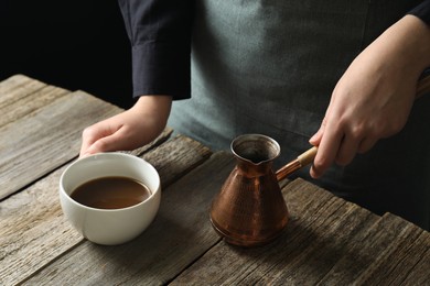 Photo of Turkish coffee. Woman with cezve and cup of freshly brewed beverage at wooden table, closeup