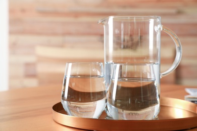 Tray with jug and glasses of water on wooden table in room, space for text. Refreshing drink