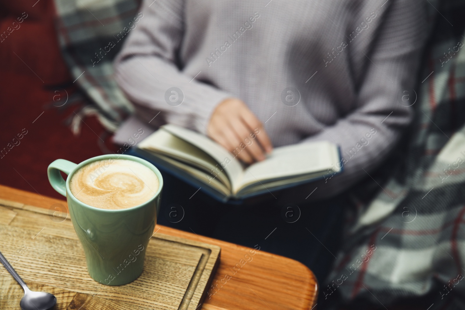 Photo of Woman reading book at table, focus on cup of coffee