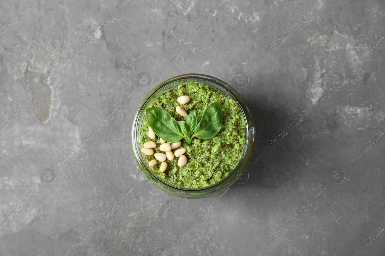 Photo of Jar with delicious pesto sauce, pine nuts and basil leaves on grey table, top view