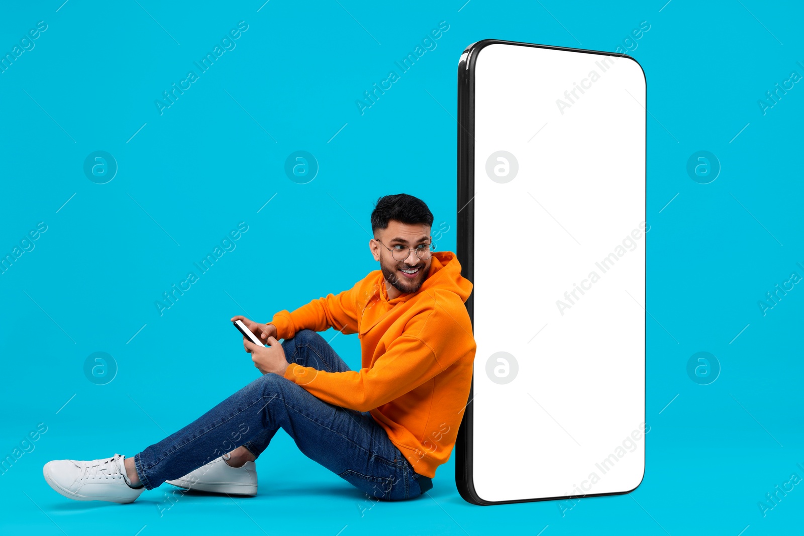 Image of Man with mobile phone sitting near huge device with empty screen on light blue background. Mockup for design