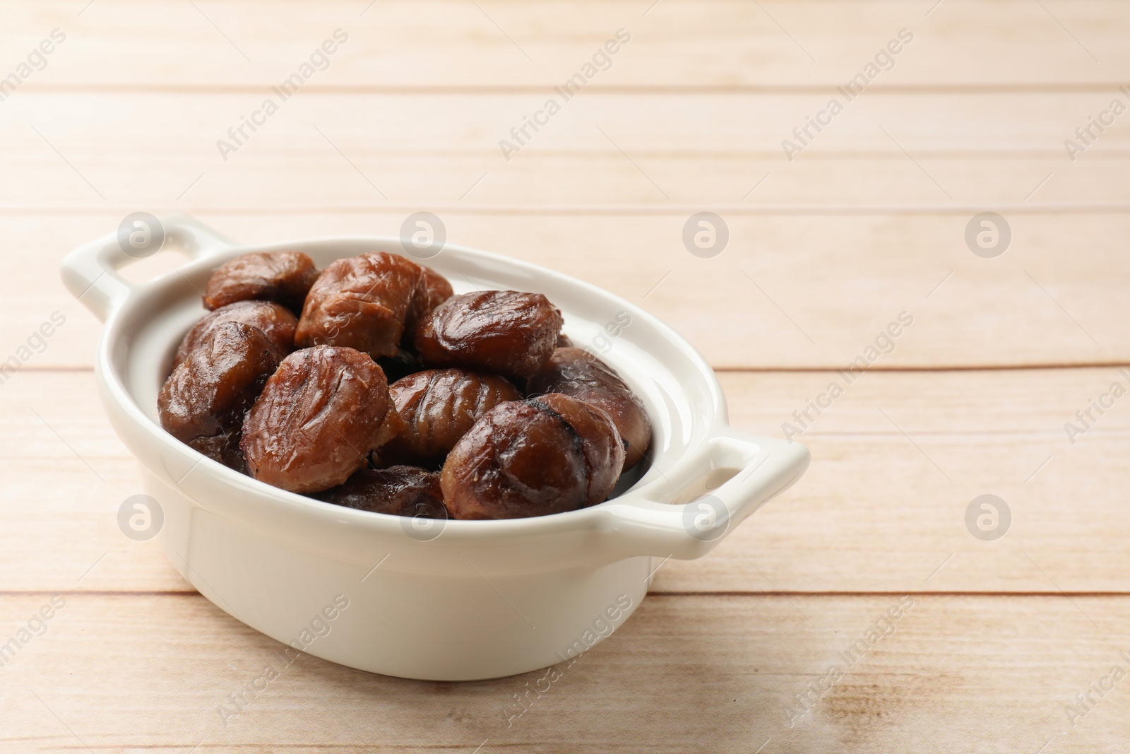 Photo of Roasted edible sweet chestnuts in dish on light wooden table, space for text