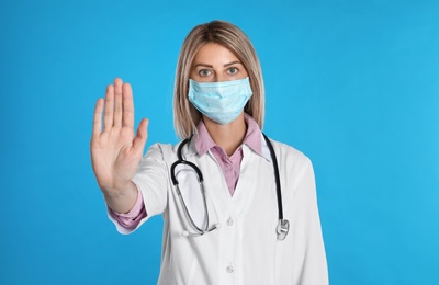 Photo of Doctor in protective mask showing stop gesture on light blue background. Prevent spreading of coronavirus