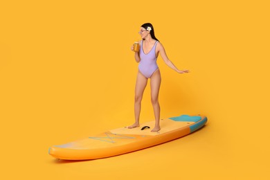 Photo of Happy woman with refreshing drink chilling on SUP board against orange background, space for text