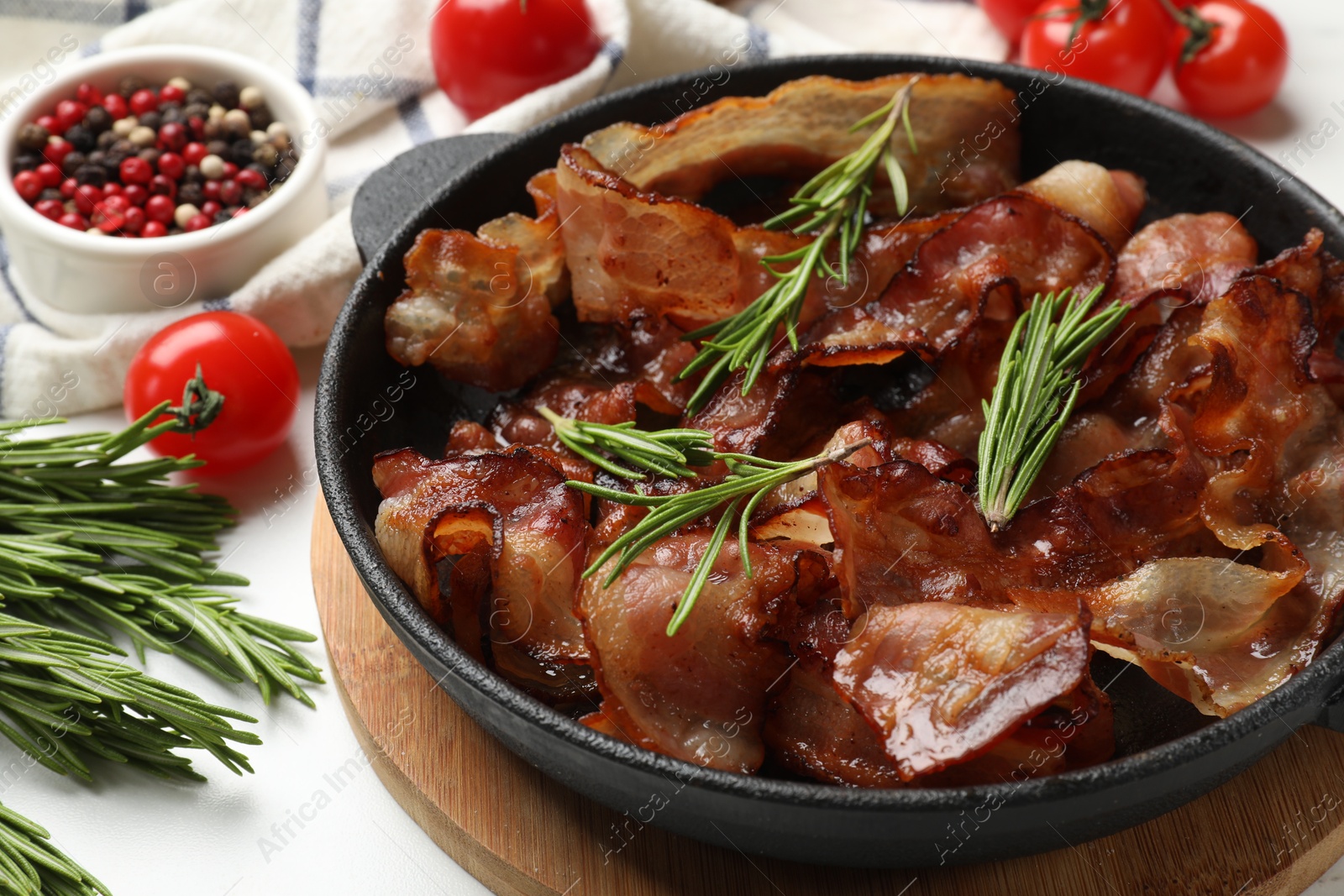 Photo of Delicious bacon slices in frying pan, spices and tomatoes on white table, closeup