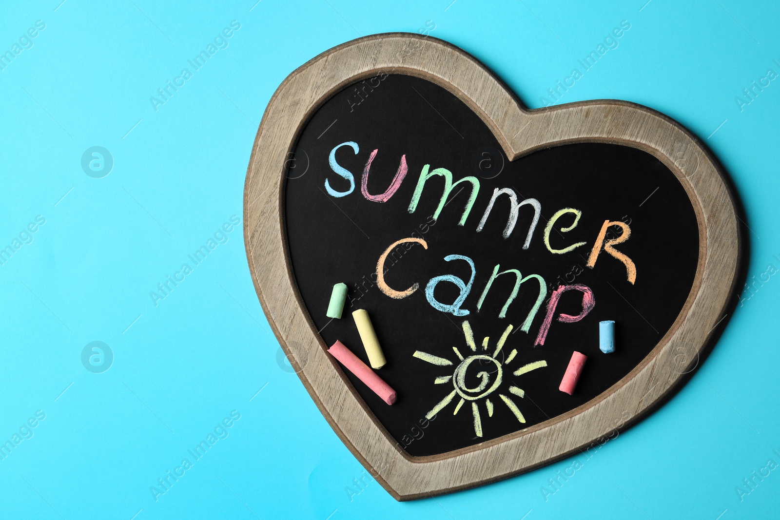 Photo of Heart shaped blackboard with text SUMMER CAMP, drawing and chalk sticks on color background, top view. Space for design