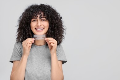 Photo of Young woman holding teeth whitening strips on light grey background, space for text