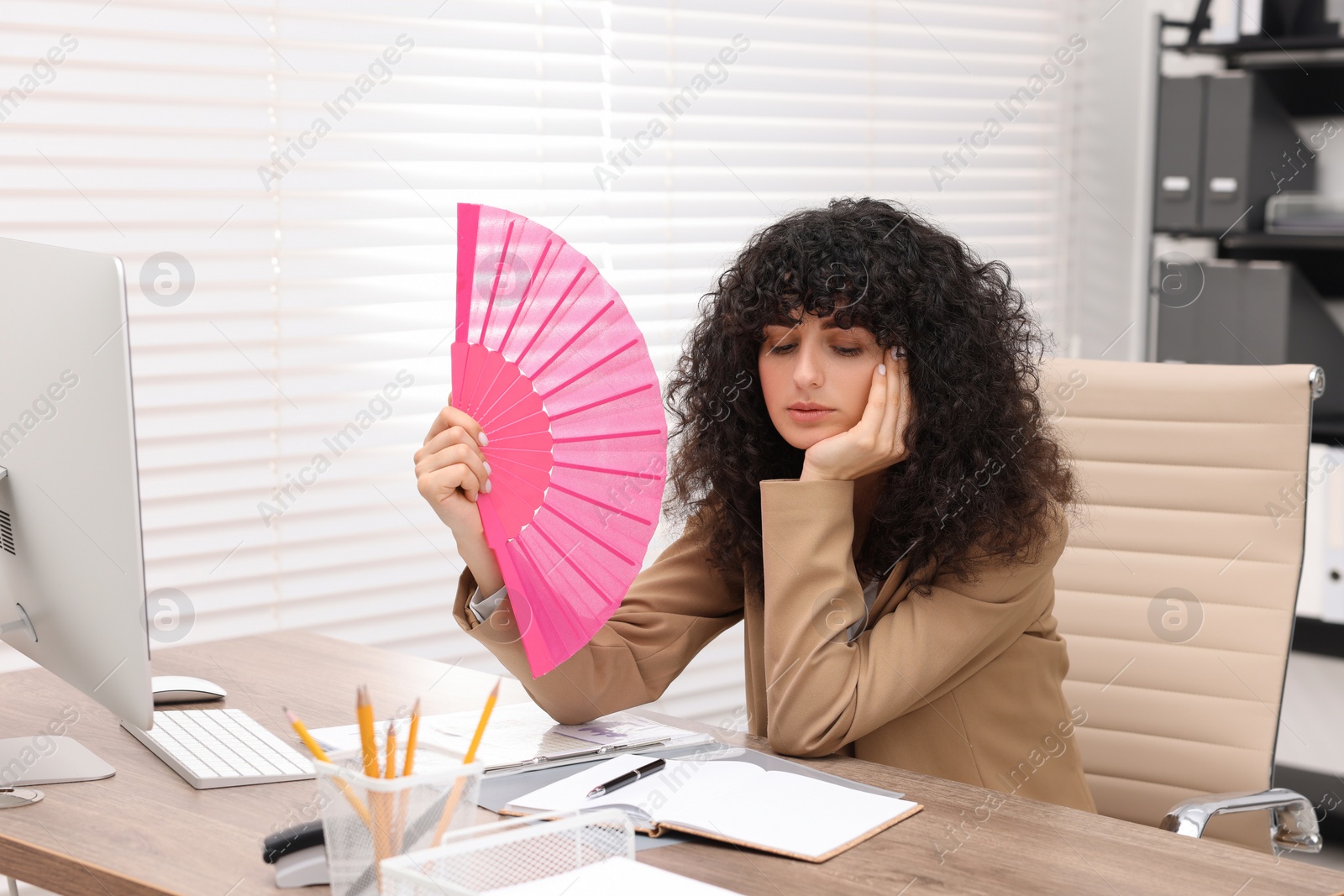 Photo of Young businesswoman waving hand fan to cool herself at table in office