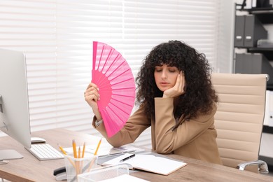 Young businesswoman waving hand fan to cool herself at table in office