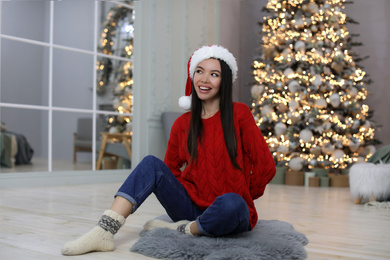 Happy young woman wearing Santa hat in room with Christmas tree