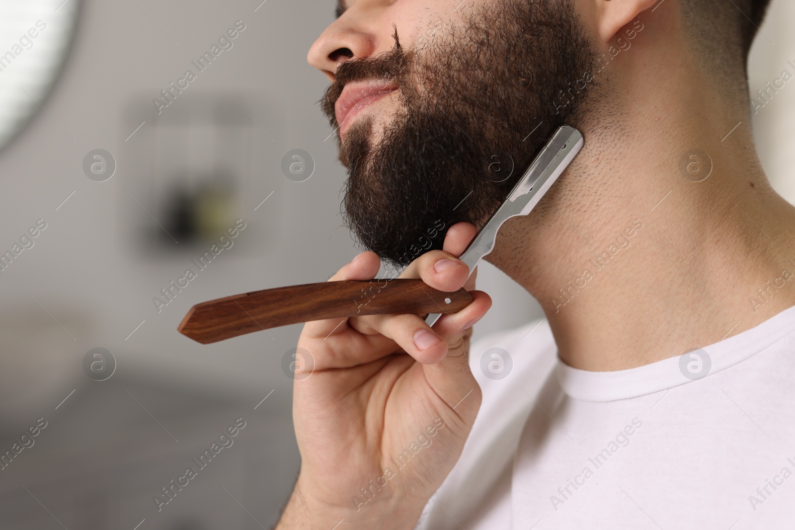 Photo of Handsome young man shaving beard with blade indoors, closeup