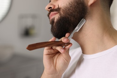 Handsome young man shaving beard with blade indoors, closeup