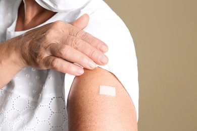 Mature woman in protective mask showing arm with bandage after vaccination on beige background, closeup
