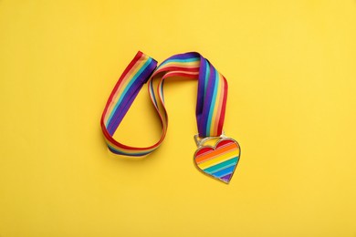 Photo of Rainbow ribbon with heart pendant on yellow background, top view. LGBT pride