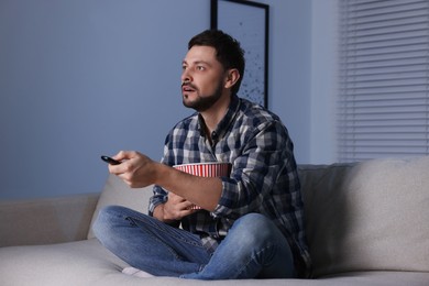 Photo of Man with popcorn bucket changing TV channels with remote control at home in evening