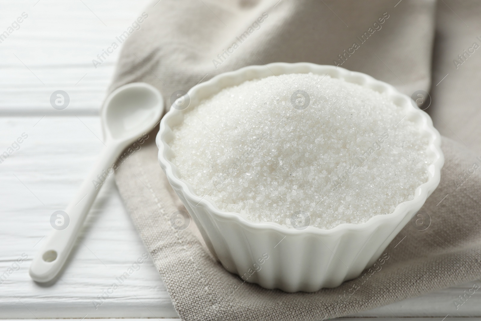 Photo of Granulated sugar in bowl and spoon on white wooden table, closeup