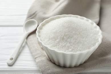 Granulated sugar in bowl and spoon on white wooden table, closeup
