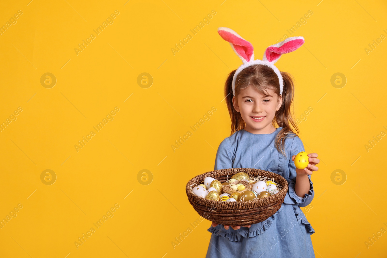 Photo of Happy little girl with bunny ears holding wicker basket full of Easter eggs on orange background. Space for text