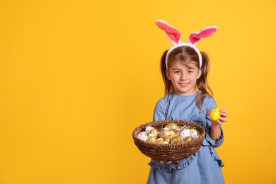 Photo of Happy little girl with bunny ears holding wicker basket full of Easter eggs on orange background. Space for text