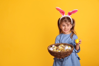 Happy little girl with bunny ears holding wicker basket full of Easter eggs on orange background. Space for text