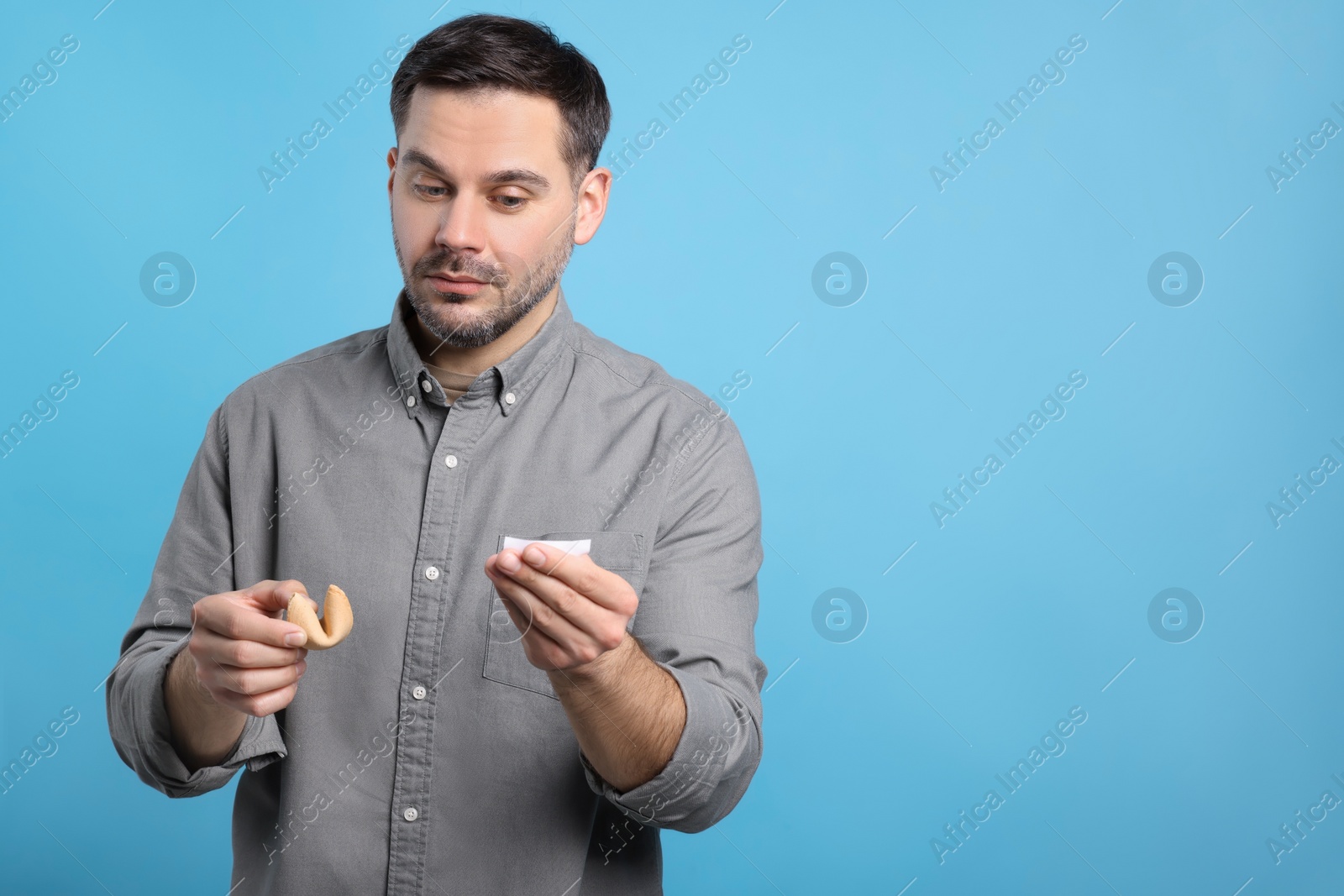 Photo of Handsome man holding tasty fortune cookie and reading prediction on light blue background. Space for text